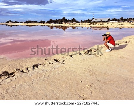 Latin adult photographer man with shorts, red shirt and hat takes pictures on the sand next to the pink colored lagoon with a high concentration of salt, Las Coloradas in Yucatan Mexico
