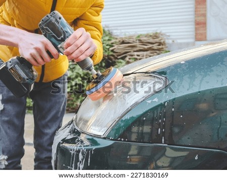 The hands of a young caucasian guy are drilling a drill with a disk with a sponge and polishing the headlight of his car with a detergent soap, close-up side view.The concept of a home car wash