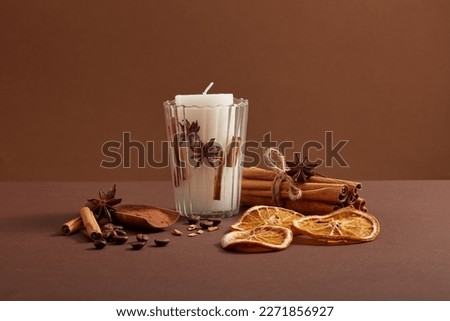 A white candle putted on a transparent glass and a bunch of cinnamon sticks, coffee beans, star anise and dried orange slices decorated around. Royalty-Free Stock Photo #2271856927