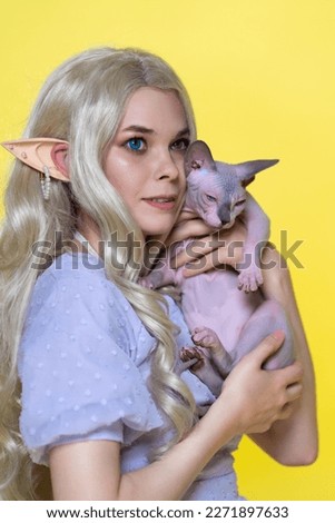 Cosplayer elf young blonde woman in blue dress with curly long hair and pearls in her ear hugging Sphinx cat to her chest, smiles, and looking away with eyes of different colors. Yellow background.