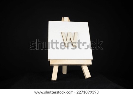 W wooden capital letter and white blank painting canvas resting on an artists easel isolated on a black background