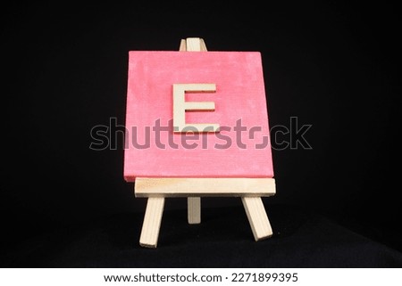 E wooden capital letter and pink blank painting canvas resting on a miniature artists easel isolated on a black background