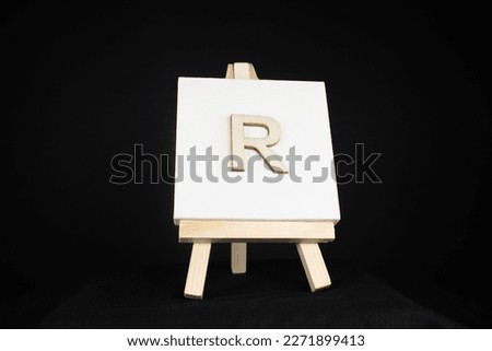 R wooden capital letter and white blank painting canvas resting on an artists easel isolated on a black background