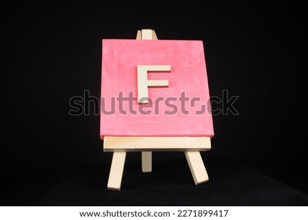 F wooden capital letter and pink blank painting canvas resting on a miniature artists easel isolated on a black background