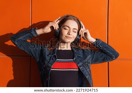 Portrait young beautiful caucasian woman leaning against wall, listening to music with headphones, eyes closed - serene, enjoying against backdrop of an orange wall in a denim jacket. Music concept