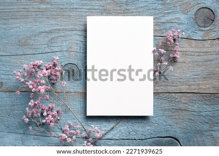 Card mockup, white blank wedding invitation with floral decor on bue wooden background. Greeting card mockup with pink dried flowers on table