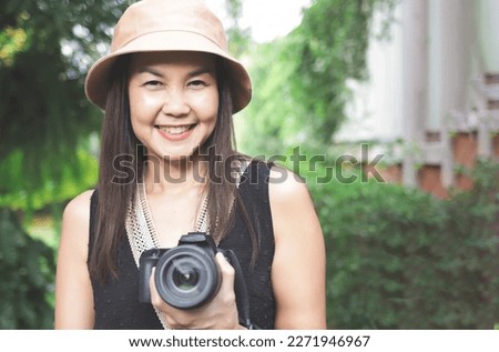 Portrait of Asian woman, wearing hat and black top sleeveless, standing in the garden and  holding dslr camera, smiling happily.