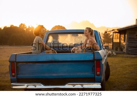 Two Women Riding In Back Of Pick Up Truck As Friends Arrive At Countryside Cabin On Road Trip Royalty-Free Stock Photo #2271977325