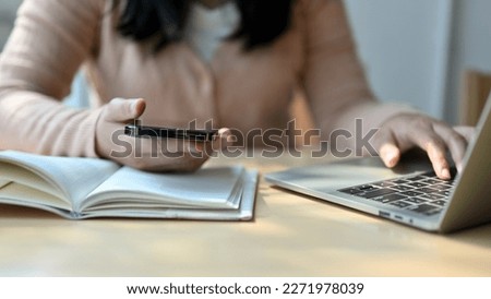 Cropped image of a young Asian female college student in casual clothes holding her phone and using her laptop to manage her schoolwork in the coffee shop or library.