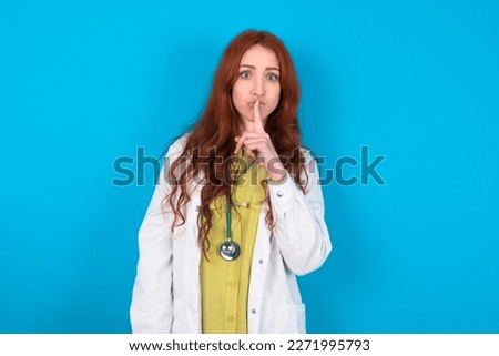 Surprised young red-haired doctor woman wearing medical uniform over blue studio background makes silence gesture, keeps finger over lips and looks mysterious at camera