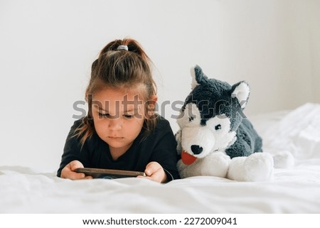 preschool age girl watching cartoons on smartphone on bed. Plush toy dog