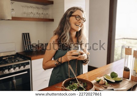Happy vegan woman smiling while holding a glass of green juice in her kitchen. Mature woman serving herself organic food at home. Woman taking care of her aging body with a healthy plant-based diet. Royalty-Free Stock Photo #2272027861