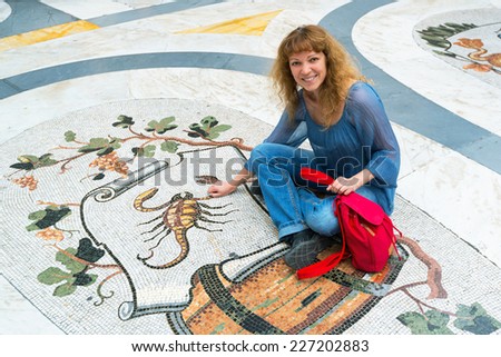Young woman sits on mosaic image of Zodiac sign Scorpio in Galleria Umberto I, Naples, Italy. Happy tourist adult girl on luxury marble floor. Concept of Astrology, travel, tourism and horoscope.