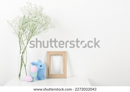 Blank frame, vase with gypsophila flowers, Easter eggs and bunny on white background