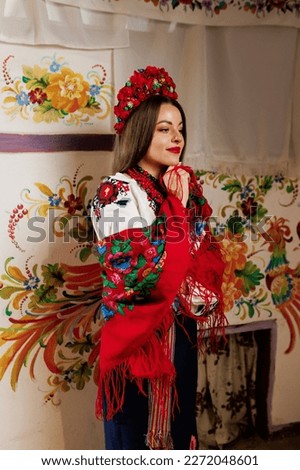 Ukrainian woman in traditional ethnic clothing and floral red wreath on  background of decorated stove in hut. Ukrainian national embroidered dress call vyshyvanka. Pray for Ukraine