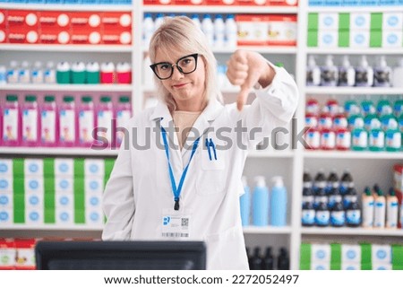 Young caucasian woman working at pharmacy drugstore looking unhappy and angry showing rejection and negative with thumbs down gesture. bad expression. 