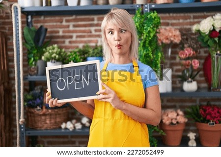 Young caucasian woman working at florist holding open sign making fish face with mouth and squinting eyes, crazy and comical. 