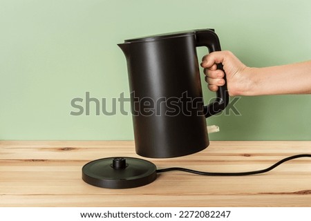 Matte black kettle kettle in a woman hand. Person hand taking off electric kettle from power base on a wooden countertop. Class 1 electrical appliance for heating water for tea and coffee. Front view.