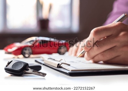 Man signing car insurance document or lease paper. Writing signature on contract or agreement. Buying or selling new or used vehicle. Car keys on table on red car background. Warranty or guarantee. Royalty-Free Stock Photo #2272152267