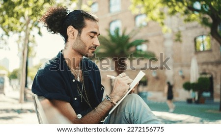 Young italian guy with ponytail and stubble sits on street bench and makes sketches with pen on piece of paper Royalty-Free Stock Photo #2272156775