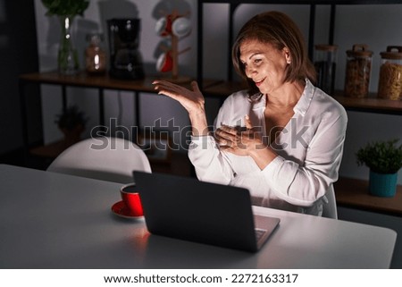 Middle age hispanic woman using laptop at home at night smiling cheerful presenting and pointing with palm of hand looking at the camera. 