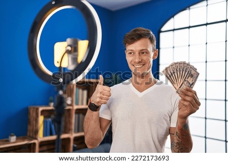Caucasian man recording vlog tutorial with smartphone at home holding money smiling happy and positive, thumb up doing excellent and approval sign 