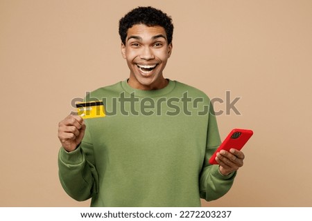 Young man of African American ethnicity wear green sweatshirt using mobile cell phone credit bank card doing online shopping order delivery book tour isolated on plain pastel light beige background Royalty-Free Stock Photo #2272203237