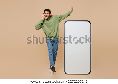 Full body young man of African American ethnicity wear green sweatshirt headohones liste music big huge blank screen mobile cell phone smartphone with area isolated on plain pastel beige background