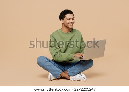 Full body young man of African American ethnicity wearing green sweatshirt sitting hold use work on laptop pc computer isolated on plain pastel light beige background studio. People lifestyle concept