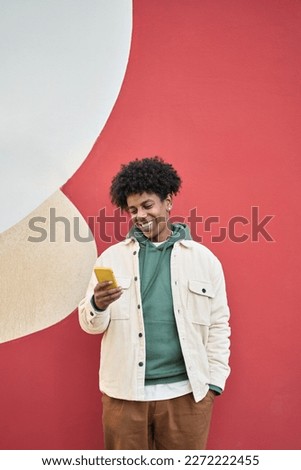Happy smiling cool gen z young African American ethnic stylish hipster guy model standing at red city urban wall outdoors using cell phone mobile device, looking at camera holding cellphone, vertical.