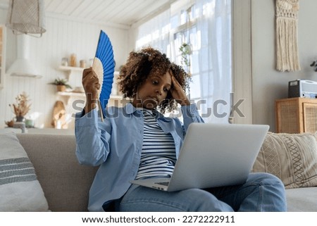 Overheating. Tired Black woman suffers from stuffiness and an inoperative air conditioner, waving blue fan sitting on couch at home working on laptop computer. High temperature, hot summer weather. Royalty-Free Stock Photo #2272222911