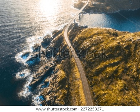 Aerial view of bridge, car, sea with waves and mountains at sunset in Lofoten Islands, Norway. Landscape with beautiful road, water, rocks and stones, golden sunlight. Top view from drone of highway