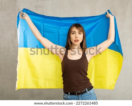 Upset patriotic young female student holding state flag of Ukraine against gray wall background indoors