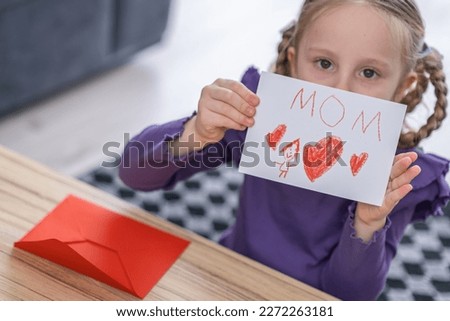 little lonely girl imagine her future mother will adopt her and take her home. She hold a drawing pitrure with letters MOM and red heart shape.
