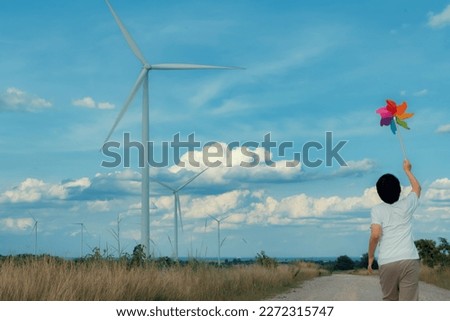 Progressive young asian boy playing with wind pinwheel toy in the wind turbine farm, green field over the hill. Green energy from renewable electric wind generator. Windmill in the countryside concept Royalty-Free Stock Photo #2272315747