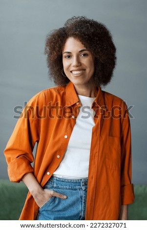 Portrait of smiling confident African American woman wearing stylish casual orange t shirt standing at home. Successful curly haired university student looking at camera, education concept