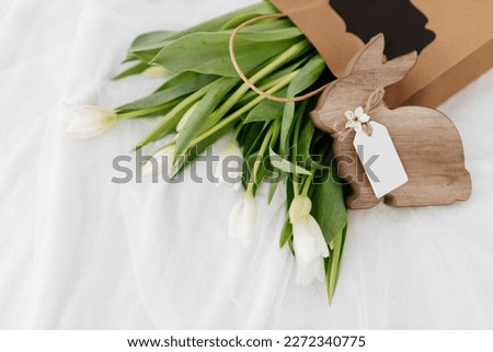 The Wooden Easter Bunny sits near a gift bag with white tulips on a light white background. Spring Background for Easter. International Women's Day, March 8th