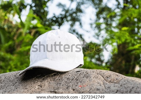 The pristine white color of this cap hat enhances the overall aesthetic of the peaceful forest setting, tastefully positioned above a nearby rock, white blank cap hat mockup image