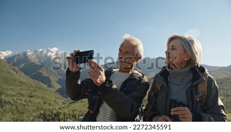 Old couple having a hike in spring mountains, then stopping to take a picture on a smartphone. Senior caucasian family spending time together travelling after retirement - tourism concept 