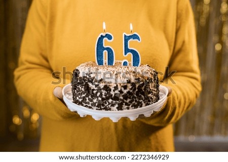 Woman holding a festive cake with number 65 candles while celebrating birthday party. Birthday holiday party people concept. Close-up view Royalty-Free Stock Photo #2272346929