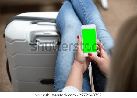 A young girl holds a mobile phone with a green screen in her hand at the airport in white sandals on a suitcase waiting for a flight.