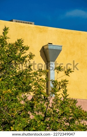 Yellow stucco cement wall with gray metal rain gutter and front yard trees with hazy dark blue sky background. Adobe style architecture in urban or suburban part of city neighborhood downtown.