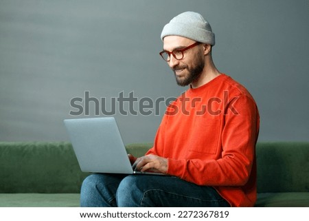 Smiling confident man programmer using laptop computer, typing on keyboard working freelance project sitting at workplace. Modern bearded hipster watching training courses, planning startup in office 