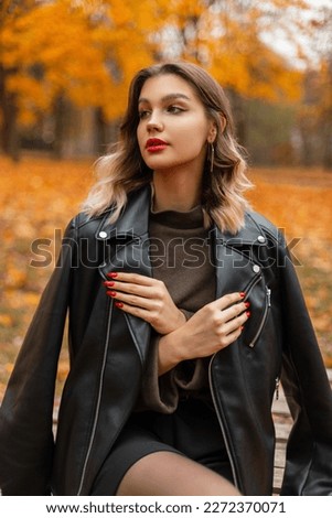 Stylish beautiful young girl with red lips in a fashionable black leather jacket and green sweater sits in an autumn park with bright yellow foliage Royalty-Free Stock Photo #2272370071