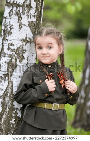 Portrait of a cute little girl in uniform with two pigtails on a green background. Victory Day, May 9 holiday.