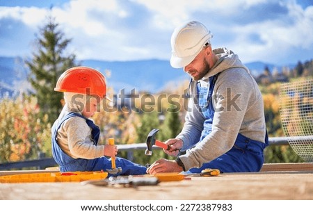 Father with toddler son building wooden frame house. Male builders hammering nail into plank on construction site, wearing helmet and blue overalls on sunny day. Carpentry and family concept. Royalty-Free Stock Photo #2272387983