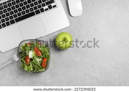 Fresh vegetable salad, apple, fork and laptop on light grey table at workplace, flat lay. Business lunch