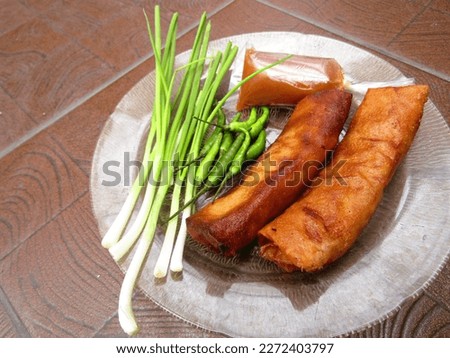 Photo of Delicious Fried Spring Rolls Objects in Golden Brown Color Along with Scallions and Fresh Green Cayenne Pepper Served on a Transparent Glass Plate with Dark Brown Background. JPG or PNG Photo