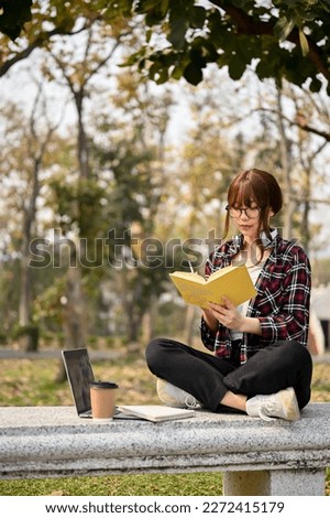 Attractive young Asian female college student in casual clothes focusing on reading a book on a bench in the campus park. leisure and lifestyle concept