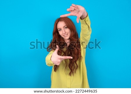 young beautiful red haired woman wearing green shirt over blue studio background making finger frame with hands. Creativity and photography concept.